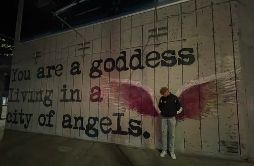 Ein Junge posiert vor einer Wand mit Street Art mit roten Engelsflügeln und einem Spruch daneben