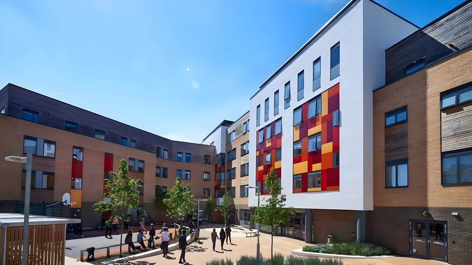 Das moderne mehrstöckige Gebäude mit bunten geometrischen Mustern zeigt das Abbey College Cambridge im Vordergrund sind Bäume und Menschen zu sehen.