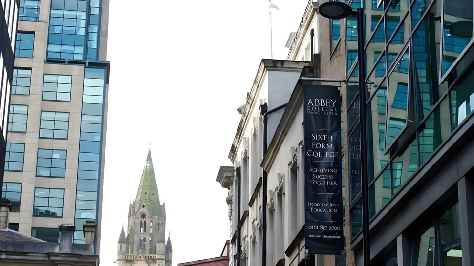 Das moderne Glas- und Stahlgebäude des Abbey College in Manchester mit einer hohen Kirchturmspitze im Hintergrund.