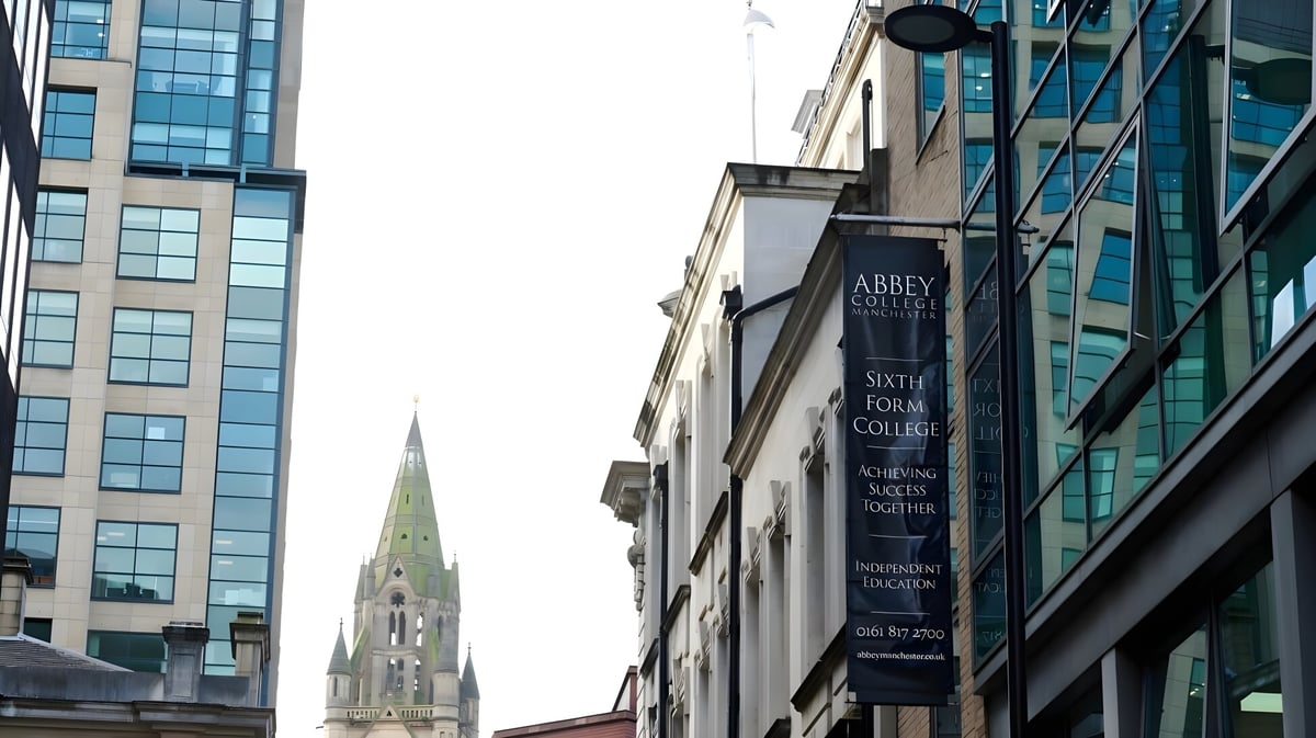 Das moderne Glas- und Stahlgebäude des Abbey College in Manchester mit einer hohen Kirchturmspitze im Hintergrund.