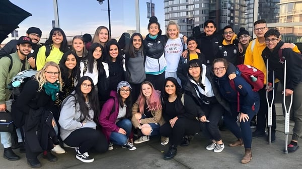 Eine Gruppe Schülerinnen steht vor hohen Gebäuden und bewölktem Himmel auf dem Gelände der Abbotsford Senior Secondary School.