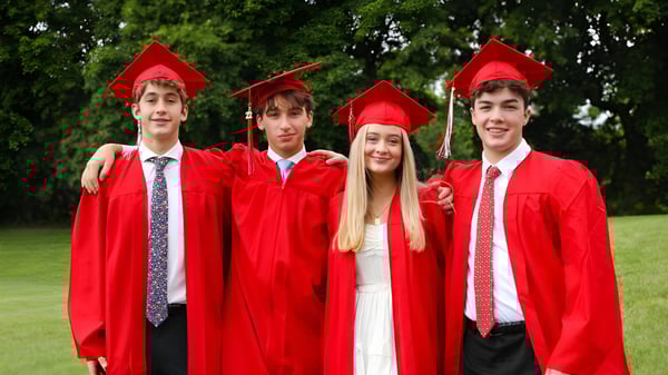 Vier Schüler der Academy of the New Church tragen rote Abschlussroben und Hüte und stehen zusammen auf einer Wiese mit Bäumen im Hintergrund.