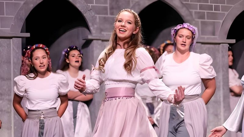 Eine Gruppe von Schülerinnen der Academy of the New Church führt in weißen und rosa Outfits auf einer Bühne mit Bogenfenstern im Hintergrund auf.