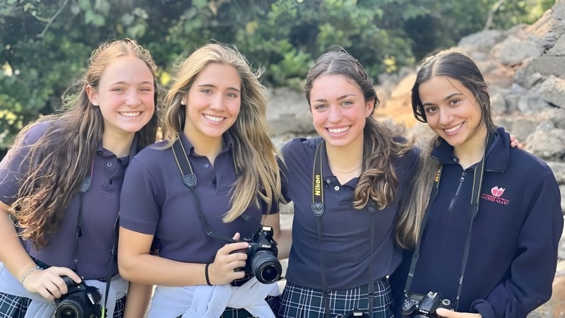 Vier Schülerinnen der Academy of the Sacred Heart stehen zusammen im Freien vor üppigem Grün.