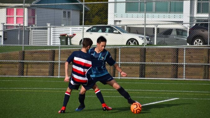 Zwei Fußballspieler kämpfen um den Ball auf dem Sportfeld des ACG Parnell College, im Hintergrund sind Wohnhäuser und Fahrzeuge zu sehen.