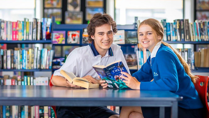 Zwei Schüler der ACG Strathallan sitzen an einem Tisch in der Bibliothek mit vielen bunten Büchern im Hintergrund.