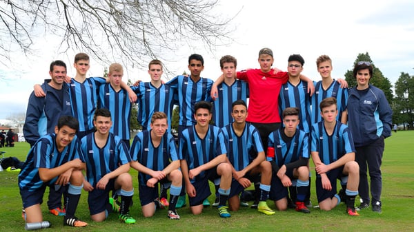 Eine Gruppe junger Fußballspielerinnen und Fußballspieler der ACG Strathallan steht zusammen auf einem Rasenfeld mit kahlen Bäumen im Hintergrund.