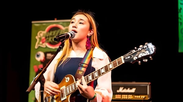 Eine Schülerin der ACG Sunderland spielt E-Gitarre und singt auf der Bühne vor einem bunten Hintergrund.