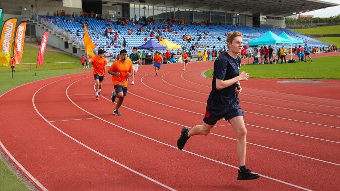 Eine Person läuft auf der Laufbahn bei einem Sportfest auf dem Campus der ACG Sunderland vor bunten Zelten und vielen Zuschauern.