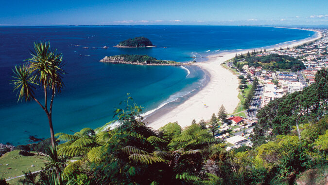 Eine Küstenlandschaft mit einem halbmondförmigen Strand und türkisfarbenem Wasser vor dem Hintergrund der Stadt ACG Tauranga.