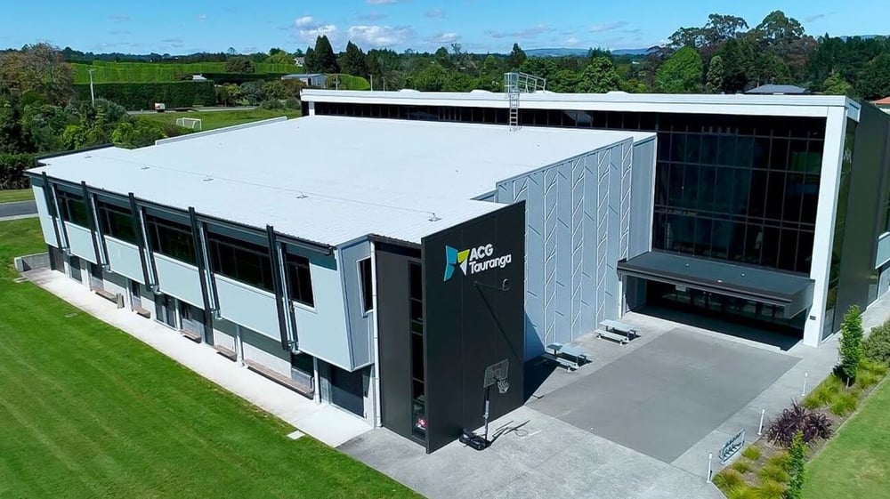 Das moderne Schulgebäude der ACG Tauranga steht auf einem grasbewachsenen Feld mit Bäumen und Bergen im Hintergrund.