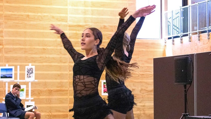 Eine Frau im schwarzen Kleid mit Federn tanzt mit erhobenen Armen vor einer Backsteinwand auf dem Campus der ACG Tauranga.