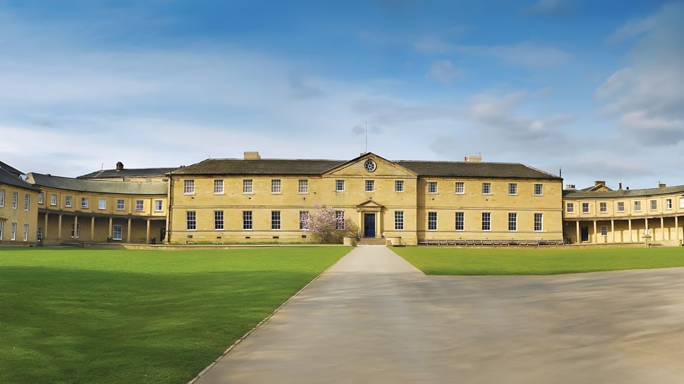 Das gelbe Hauptgebäude der Ackworth School steht auf einem großen Rasenfeld unter einem blauen Himmel mit Wolken.