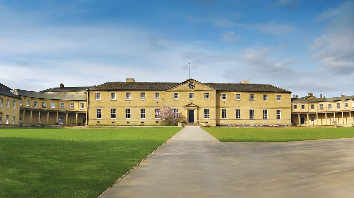 Das gelbe Hauptgebäude der Ackworth School steht auf einem großen Rasenfeld unter einem blauen Himmel mit Wolken.