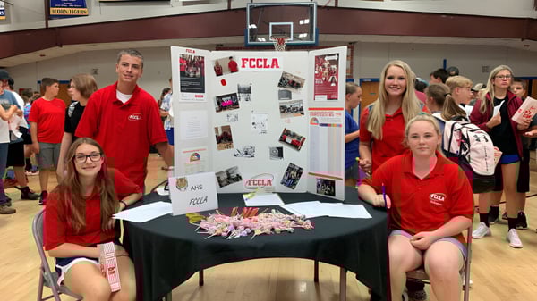 Eine Gruppe Schülerinnen und Schüler der Acorn High School steht in der Turnhalle um einen Präsentationstisch mit Postern und Materialien.