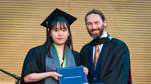 Eine Absolventin in Abschlussrobe steht mit einem Mann in akademischer Tracht und hält ein Diplom auf dem Campus der ACS International School Cobham.