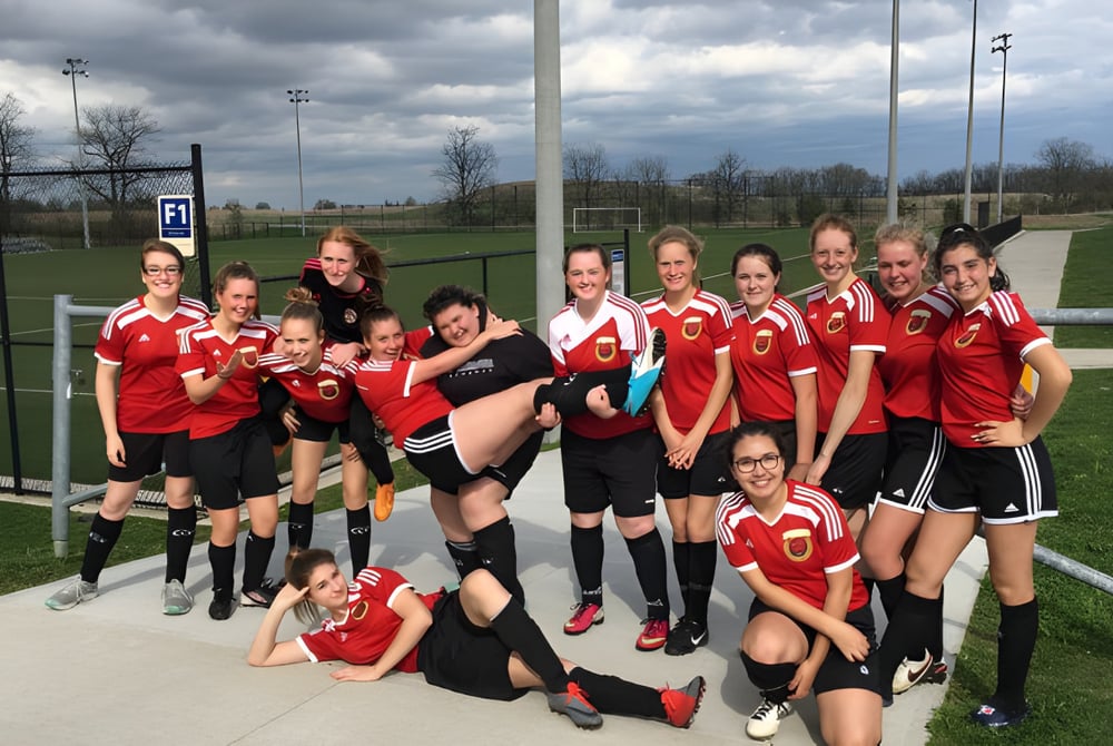 Eine Gruppe von Fußballspielerinnen der Acton High School posiert auf dem Sportfeld unter bewölktem Himmel.