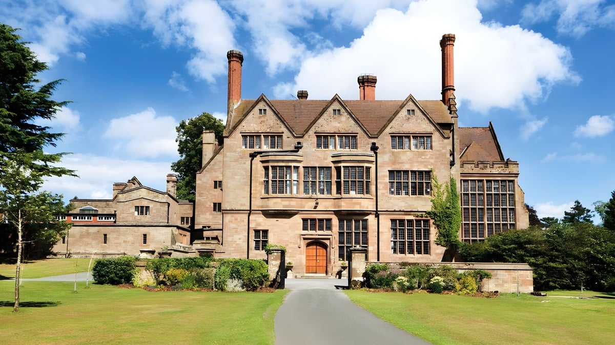Das historische Herrenhaus der Adcote School steht umgeben von gepflegten Rasenflächen und grüner Vegetation unter blauem Himmel.