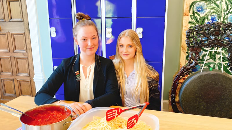 Zwei Schülerinnen der Adcote School sitzen gemeinsam an einem Tisch und essen eine Mahlzeit in einem Raum mit blauer Wand und Pflanzen.