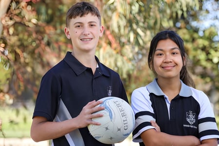 Zwei Schüler der Adelaide High School stehen gemeinsam im Freien und halten einen Rugbyball vor Bäumen.