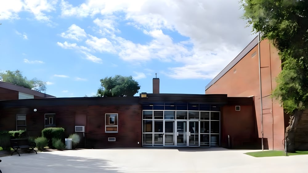 Das moderne Backsteingebäude des Aden Bowman Collegiate ist von viel Grün umgeben unter blauem Himmel mit Wolken.