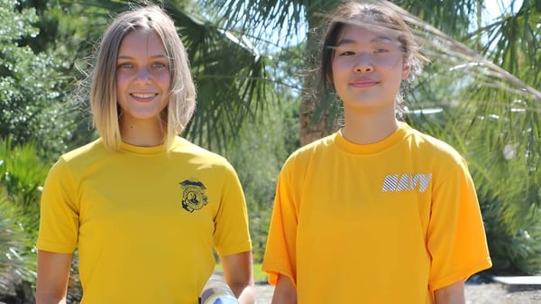 Zwei Schüler der Admiral Farragut Academy stehen mit leuchtend gelben Hemden vor Palmen im tropischen Umfeld.