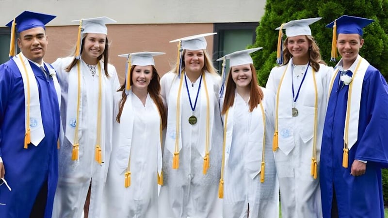 Eine Gruppe von Absolventinnen und Absolventen der Adrian High School steht in blauen und weißen Abschlussroben vor einem Gebäude und feiert ihren Abschluss.