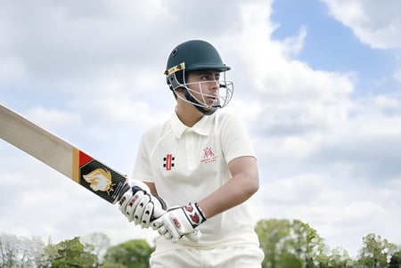 Ein Cricket-Spieler der Akeley Wood School steht mit weißer Uniform und Helm auf dem Spielfeld vor bewölktem Himmel und Bäumen.