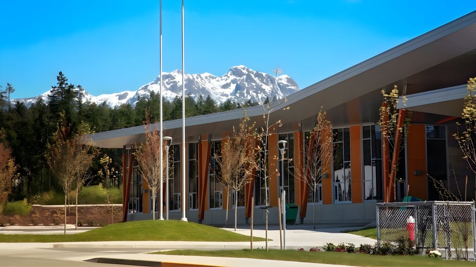 Das moderne Gebäude der Alberni District Secondary School mit großen Fenstern und einer Rasenfläche im Vordergrund vor verschneiten Bergen.