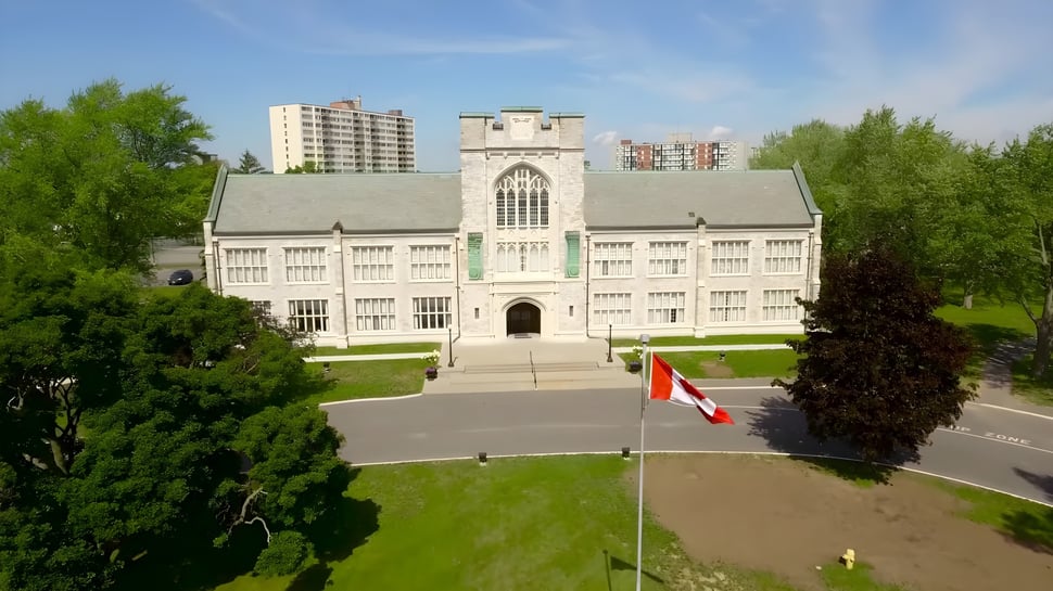 Das historische Gebäude des Albert College steht in einer gepflegten Grünanlage mit Bäumen und Hochhäusern im Hintergrund.