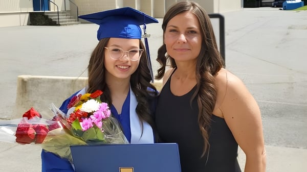 Eine Schülerin der Aldergrove Community Secondary School steht in blauer Abschlussrobe neben einer älteren Frau mit Blumen vor dem Schulgebäude.