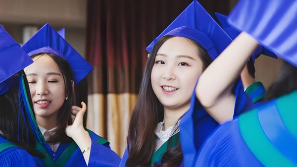 Zwei Schülerinnen der Alexander Academy feiern in blauen Abschlussroben vor einem roten Vorhang ihre Graduierung.