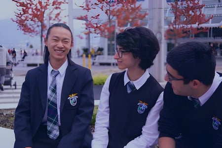 Drei Schüler in Schuluniform stehen vor einem Gebäude mit roten Bäumen auf dem Gelände der Alexander Academy.