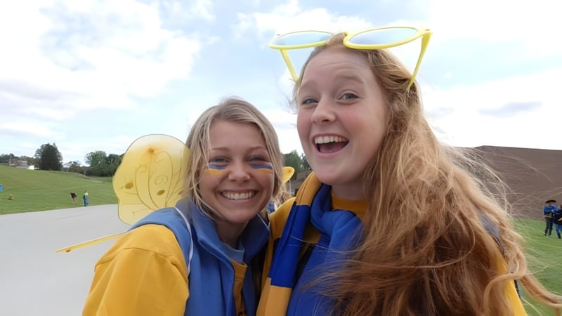 Zwei Schülerinnen der Alexander Galt Regional High School in gelben und blauen Outfits lachen im Freien auf einer Grünfläche.