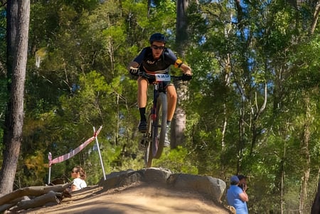 Ein Mountainbiker führt einen Sprung über eine Erdschanze auf dem Gelände der Alexandra Hills State High School aus, während Zuschauer zuschauen.