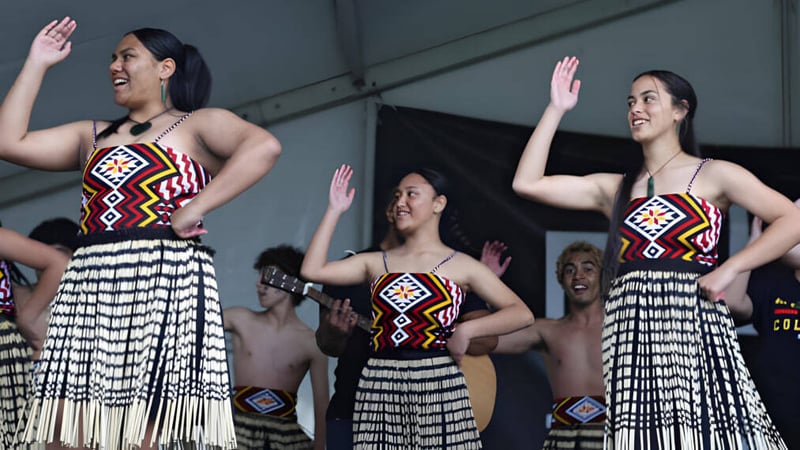 Drei Frauen führen auf der Bühne vor dem Publikum des Alfriston College einen traditionellen Tanz in farbenfrohen Kleidern auf.