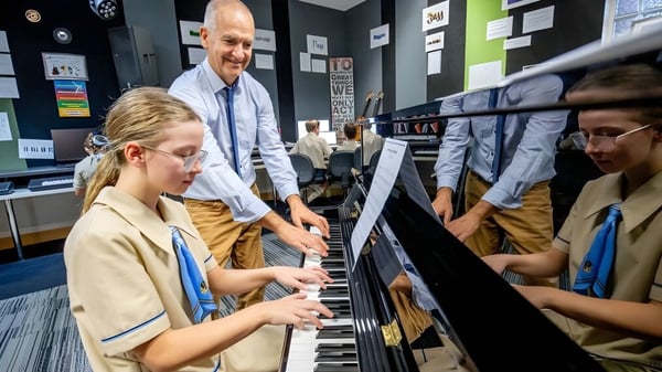 Schüler und Lehrer der All Saints Anglican School versammeln sich um ein Klavier, während eine Schülerin spielt.
