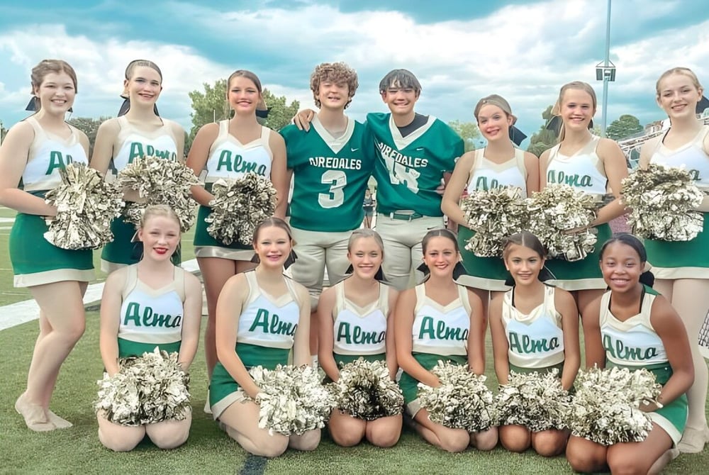 Eine Gruppe Cheerleader posiert in grünen und weißen Uniformen auf dem Sportfeld der Alma High School.
