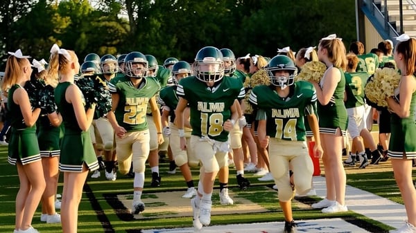 Eine Gruppe von Fußballspielern in grünen und weißen Trikots steht auf dem Sportplatz der Alma High School.