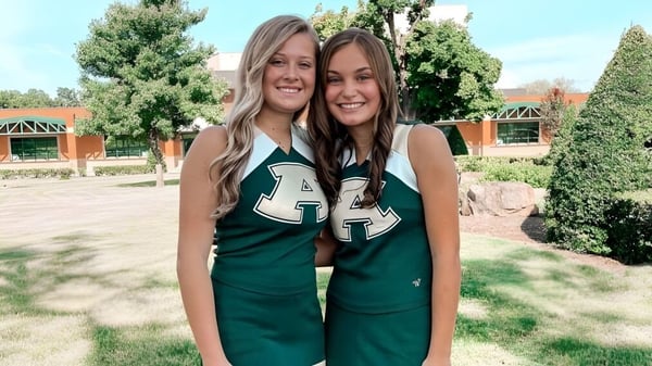 Zwei Schülerinnen der Alma High School stehen in grünen Trikots auf einer Wiese mit Bäumen und Gebäuden im Hintergrund.