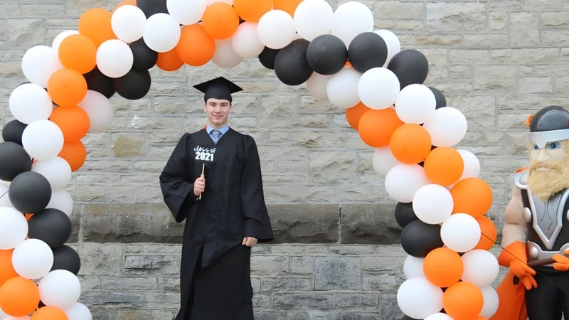 Ein Schüler der Almonte & District High School steht in einem Abschlussgewand unter einem Bogen aus bunten Luftballons vor einer Backsteinwand.