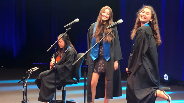 Drei Schülerinnen der Alpha School stehen in Abschlusskleidung auf der Bühne und sprechen in Mikrofone vor blau beleuchtetem Hintergrund.