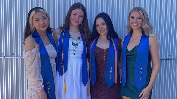 Vier Schülerinnen der Alpha School stehen in bunten Kleidern vor einer blauen Wellblechwand.