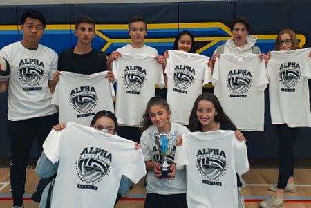 Eine Gruppe von Schülerinnen und Schülern der Alpha School posiert in weißen T-Shirts mit Alpha-Logo in einer Sporthalle mit blauen und gelben Wänden.