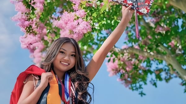 Eine junge Frau mit langen dunklen Haaren hebt lächelnd die Arme umgeben von pinken Blumen auf dem Gelände des Anaheim Union High School District.