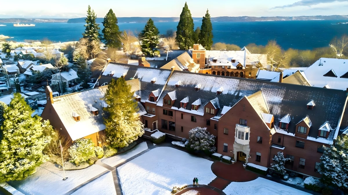 Das historische rote Backsteingebäude der Annie Wright Schools ist umgeben von schneebedeckten Bäumen und liegt nahe einem Gewässer mit Bergen im Hintergrund.