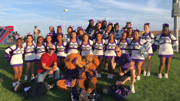 Eine Gruppe Cheerleader in lila und weißen Uniformen steht auf dem Sportfeld der Antelope Valley Union High School District vor blauem Himmel.
