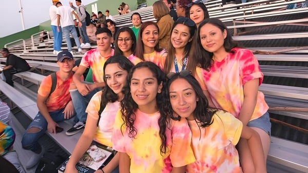 Eine Gruppe lachender Schülerinnen in bunten Tie-Dye-Shirts sitzt auf den Tribünen auf dem Campus der Antelope Valley Union High School District mit Blick auf das Sportfeld.
