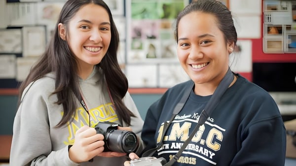Zwei Schülerinnen des Aotea College stehen in einem Innenraum, wobei eine von ihnen eine Kamera hält.