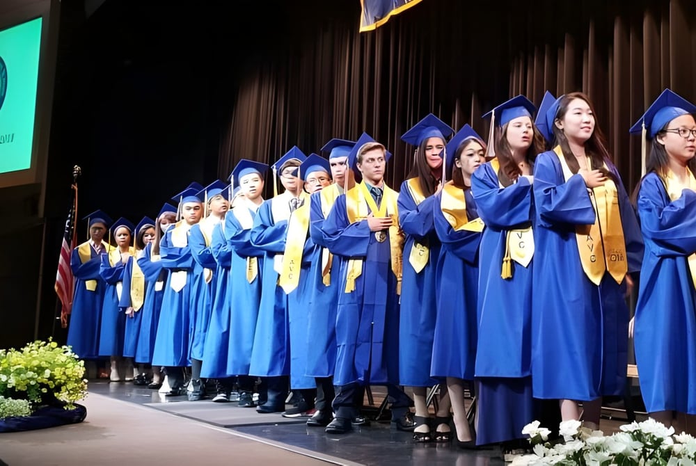 Eine Gruppe Absolventen der Apple Valley Christian School steht in blauen Talaren auf einer Bühne vor einem grünen Logo.